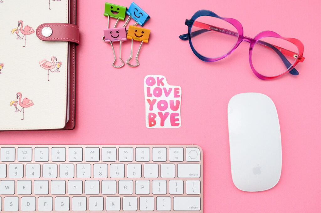 Pink desk with keyboard, mouse, glasses, flamingo notebook, binder clips, and sticker, perfect for writing client emails.